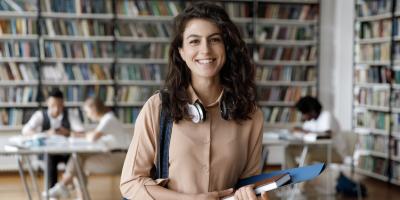 Happy Hispanic gen Z student girl with headphones visiting public library for work on study research project, holding learning papers, notebook, looking at camera, smiling.