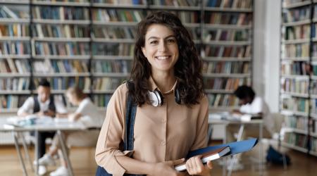 Happy Hispanic gen Z student girl with headphones visiting public library for work on study research project, holding learning papers, notebook, looking at camera, smiling.